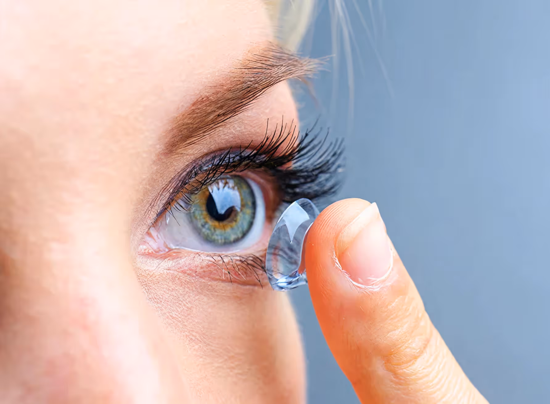 Close-up of a person holding a transparent contact lens on their fingertip near their green eye.