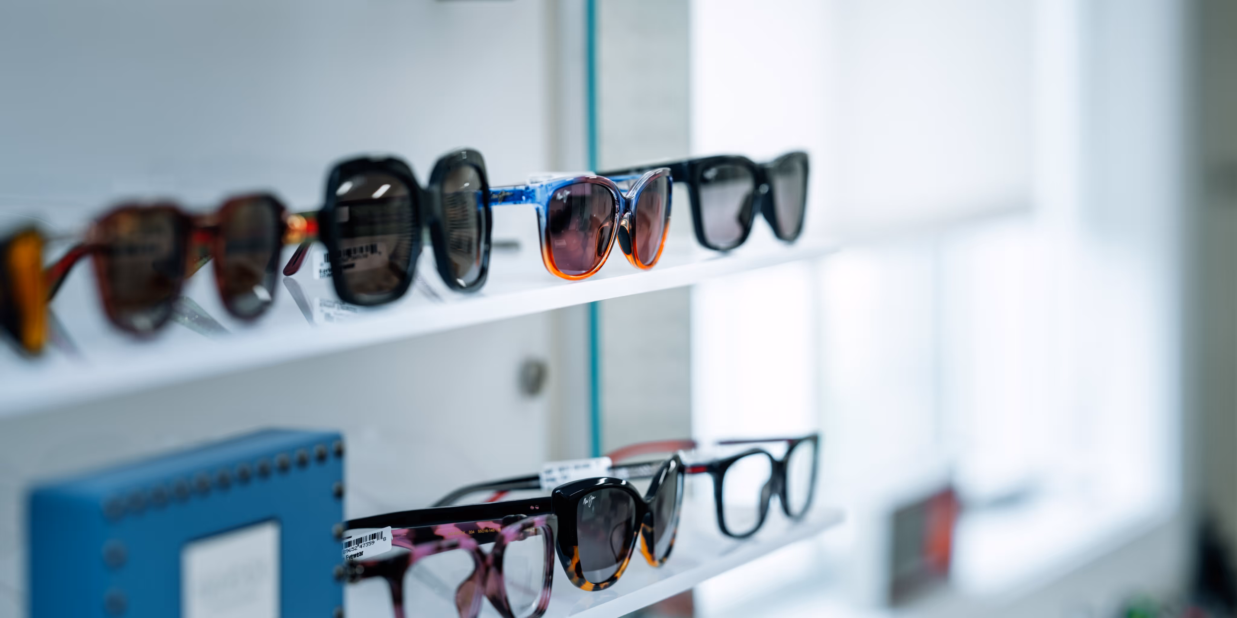 Several pairs of sunglasses with different frame styles and colors displayed on two white shelves in a bright store.