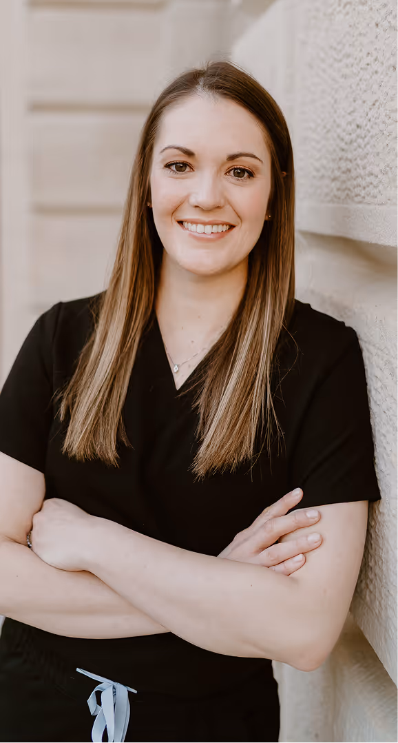 Smiling woman with long straight hair wearing black medical scrubs, standing with arms crossed against a textured wall.