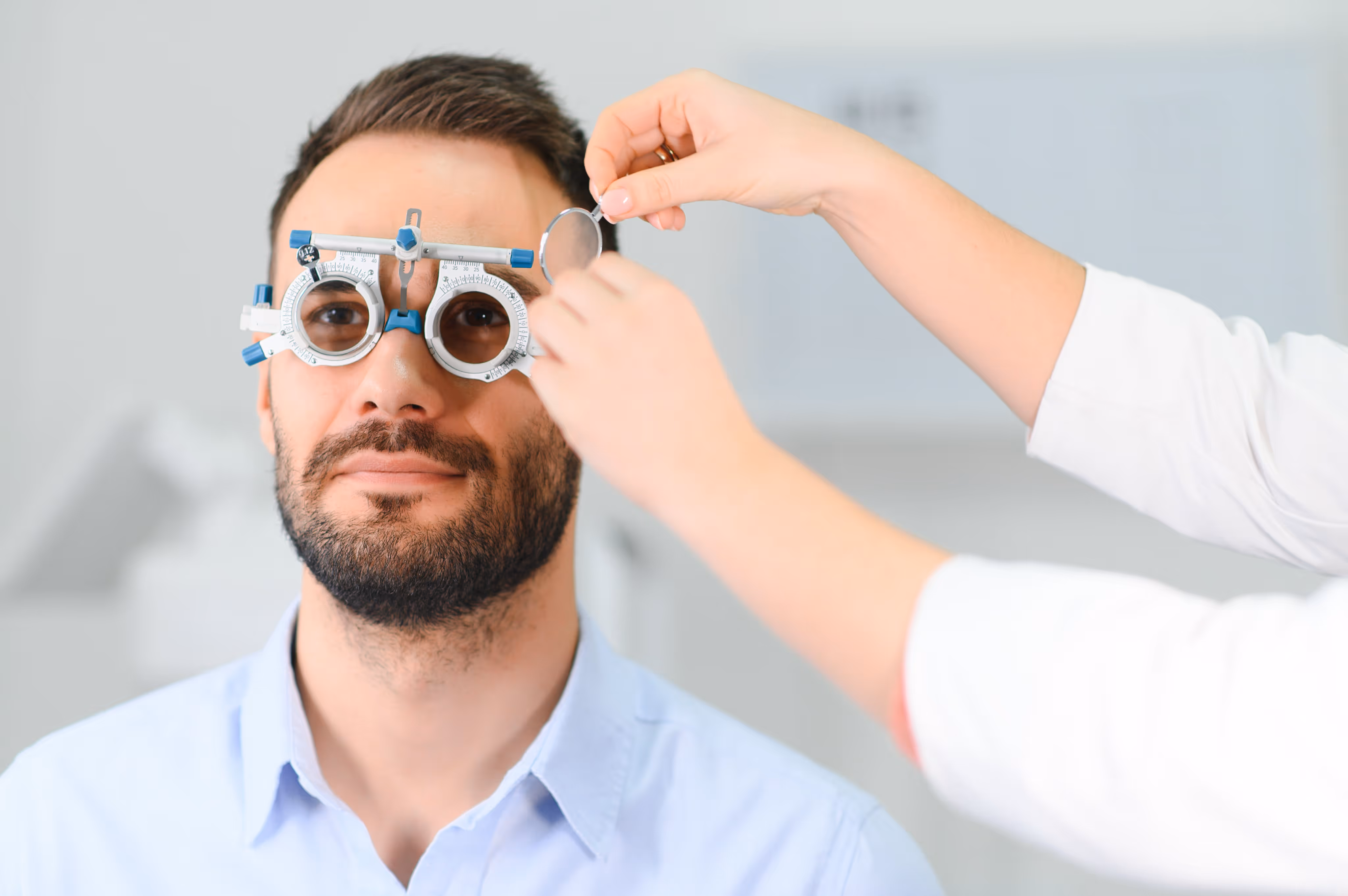 Man with beard undergoing an eye exam wearing a trial frame while a professional adjusts lenses.