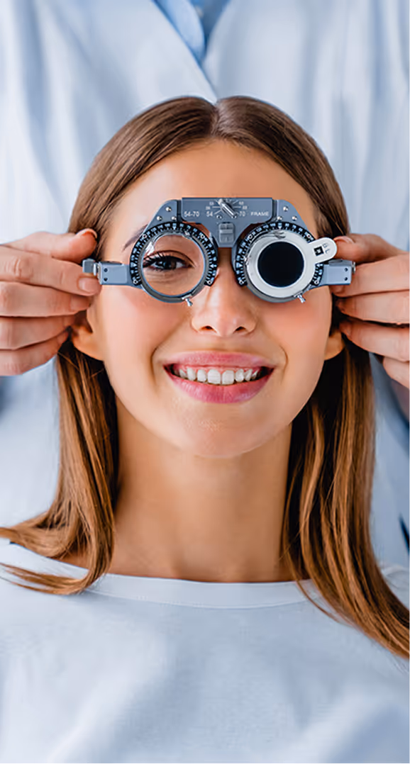 Smiling woman undergoing an eye exam with a phoropter held by a person in a white coat behind her.