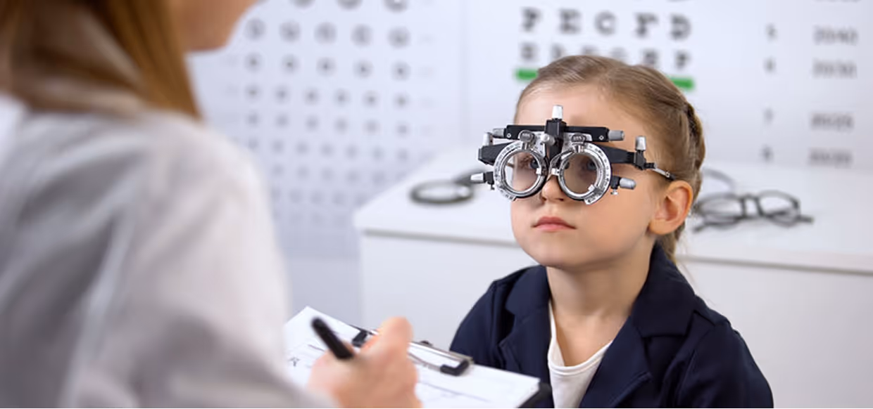 A young girl wearing a trial frame undergoes an eye exam with an optometrist holding a clipboard.