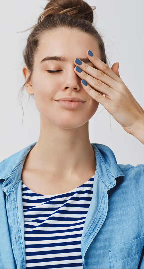 Young woman with hair in a bun, wearing a blue striped shirt and denim jacket, gently covering one eye with her hand painted with blue nail polish.