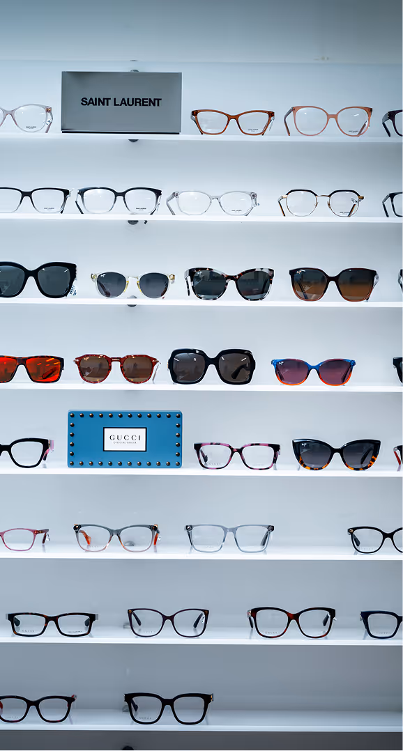 Display of various eyeglasses and sunglasses on white shelves with Saint Laurent and Gucci brand signs.
