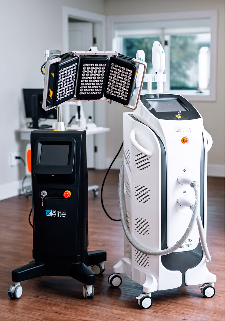 Two medical aesthetic devices on wheels, one black with LED panels and one white with a touchscreen and handpiece attachments in a clinical room.