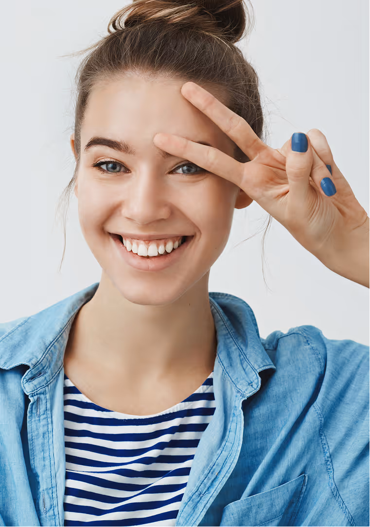 Smiling young woman with blue eyes and a top bun making a peace sign near her forehead, wearing a blue striped shirt and denim jacket.