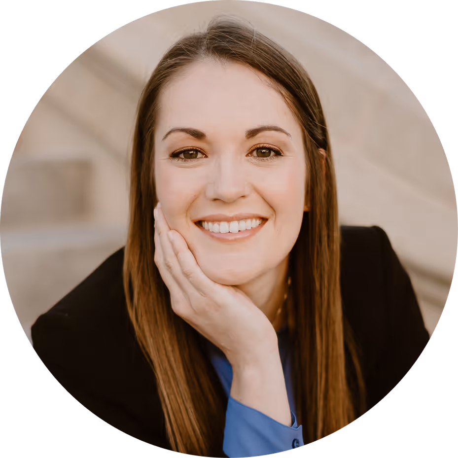 Smiling woman with long straight brown hair resting her head on her hand, wearing a black blazer and blue shirt.