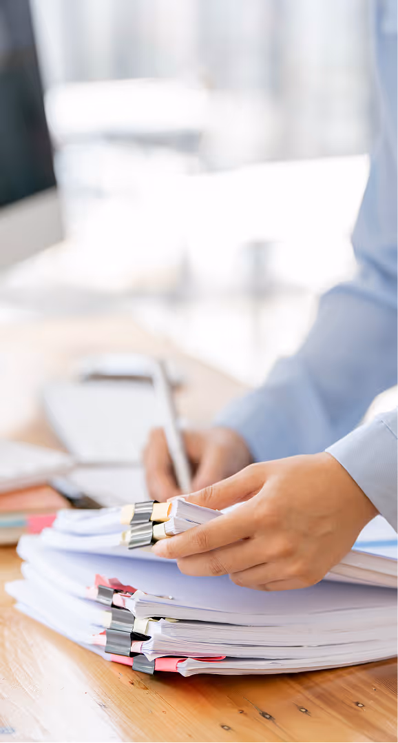 Person organizing and writing on stacks of clipped documents on a wooden desk.