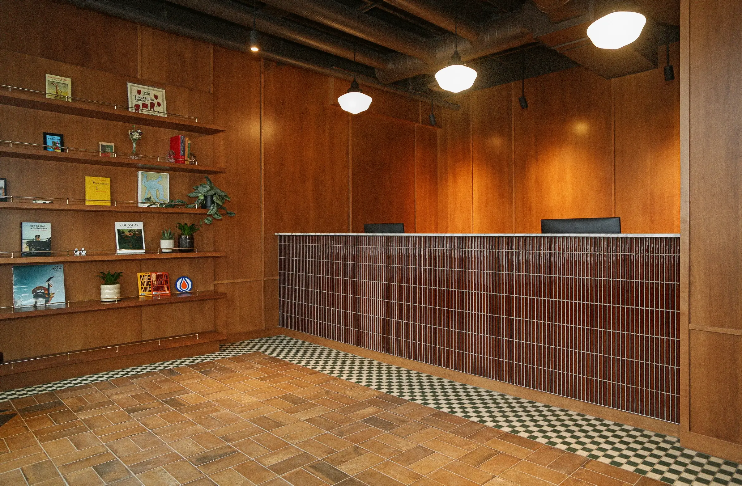 Reception area of General Dental with wooden walls, a front desk with vertical brown tiles, pendant lights, and wooden shelves holding books and plants.