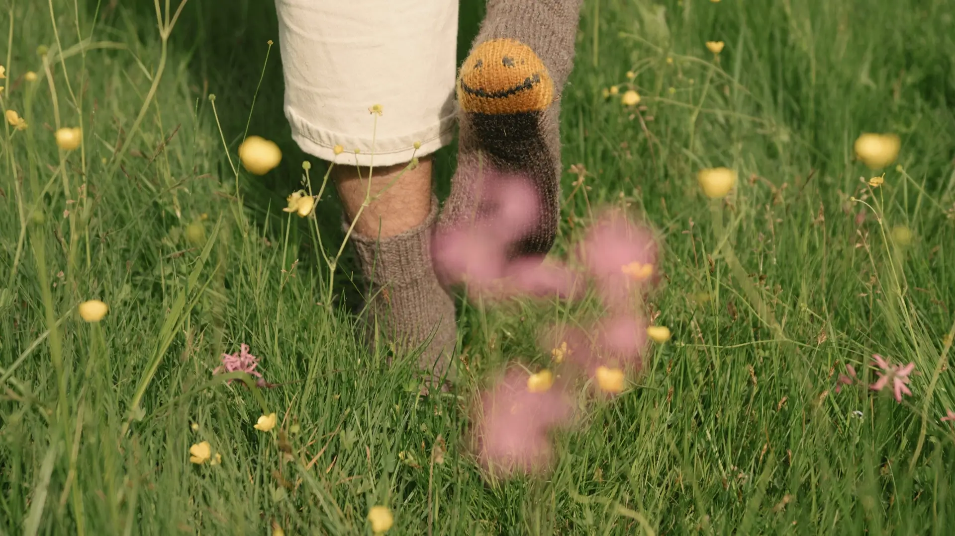 Person walking barefoot in a green meadow with yellow and pink wildflowers, wearing a sock with a smiley face hanging from one foot.