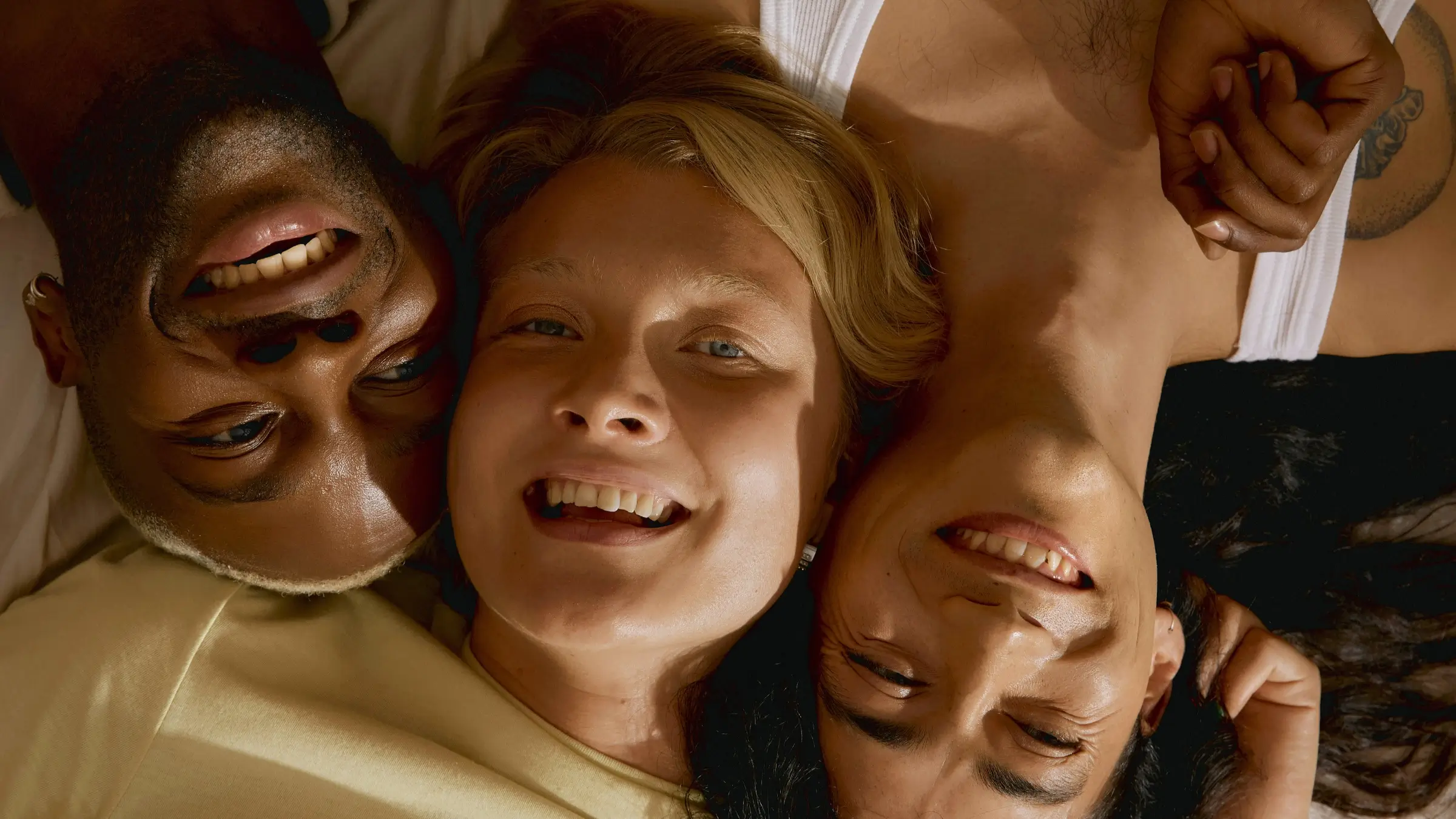 Three diverse friends lying close together on a bed, smiling and looking happy.