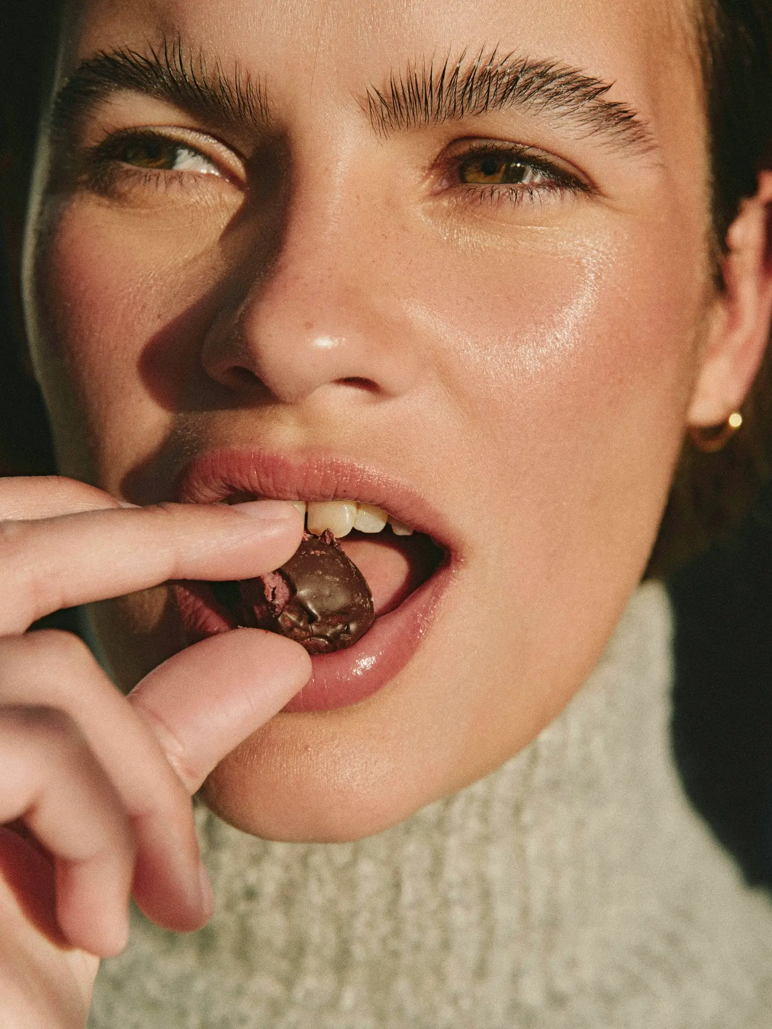 Close-up of a person with brown eyes holding a bitten chocolate piece near their lips, wearing a cream knitted sweater and a small gold hoop earring.