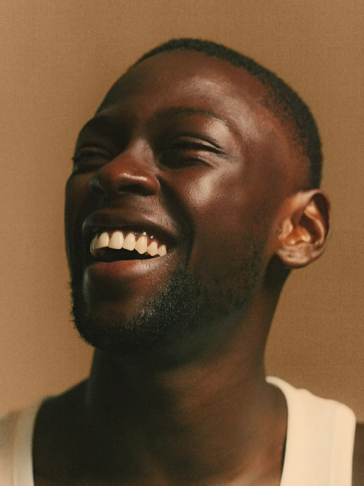 Close-up of a young man with a beard smiling and laughing against a neutral brown background.