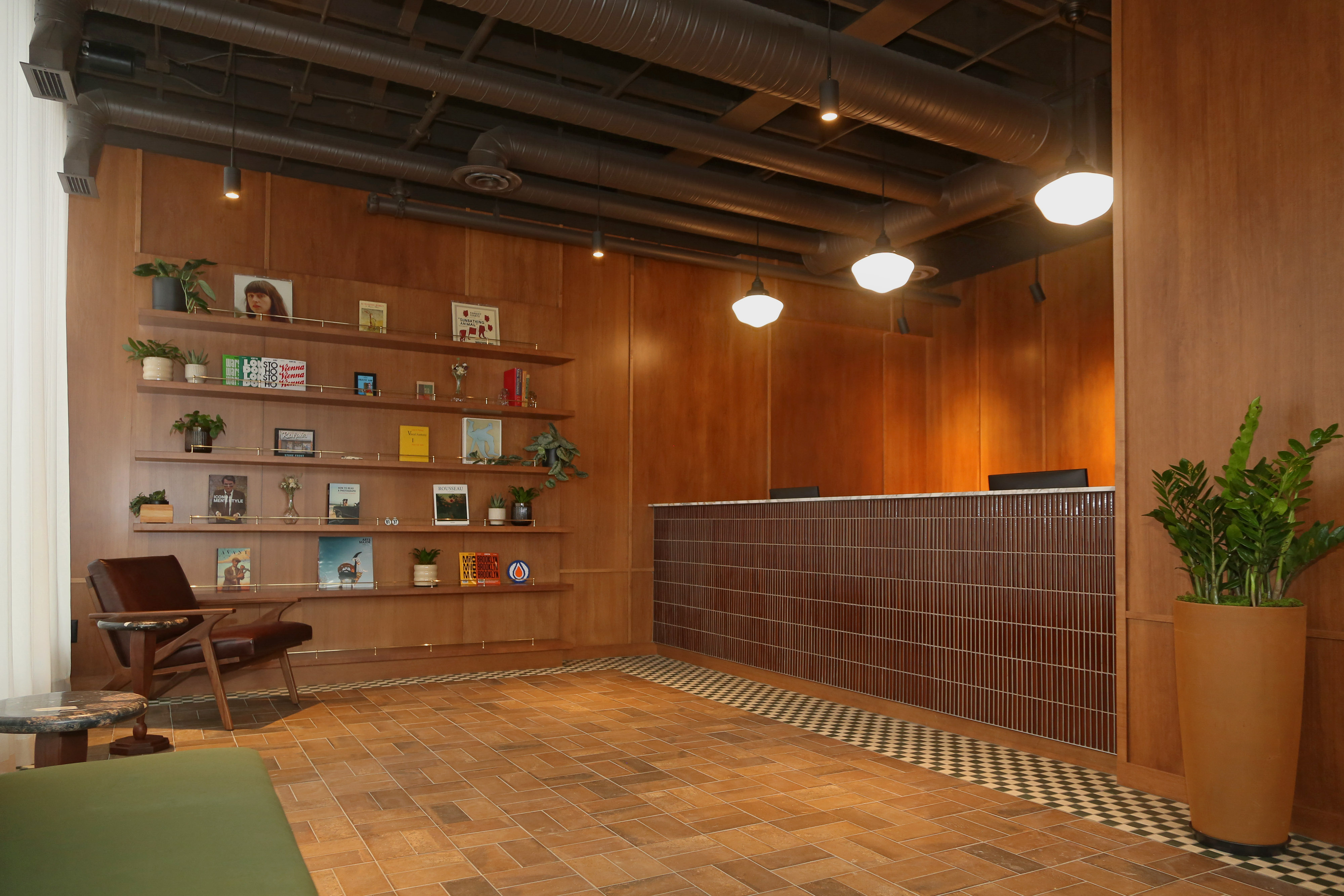 Modern lobby with wood-paneled walls, a tiled reception desk, wooden shelves displaying books and plants, a leather chair, and hanging ceiling lights.