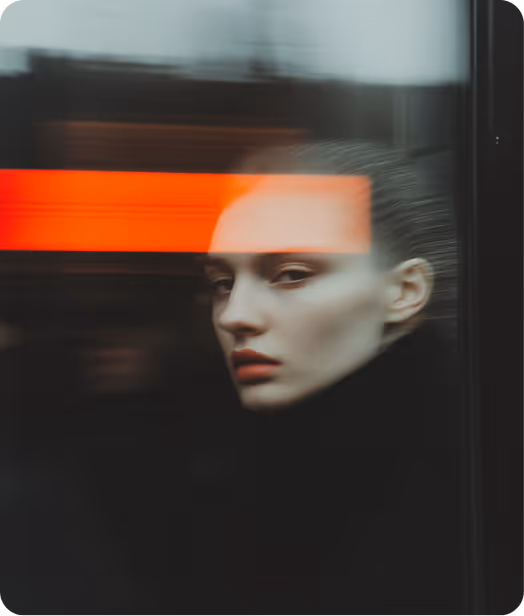 Portrait of a woman with pale skin and red lipstick looking through a window with a bright orange reflection across her forehead.