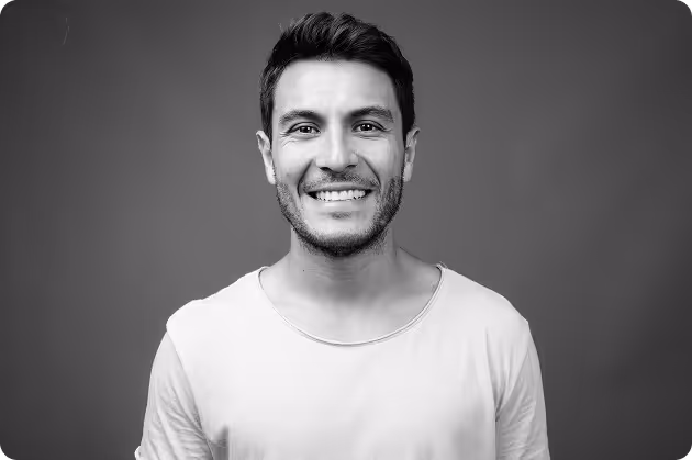 Smiling man with short dark hair and stubble wearing a plain light-colored t-shirt against a plain background.