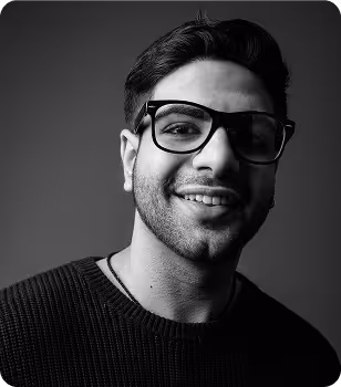 Black and white portrait of a smiling man wearing large glasses and a dark sweater against a plain background.