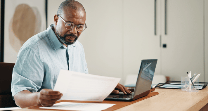 Municipal finance officer reviewing printed financial documents while working on a laptop at his desk.