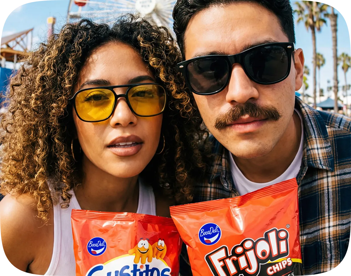 Close-up of a woman with curly hair wearing yellow-tinted sunglasses and a man with a mustache wearing black sunglasses, both holding bags of BocaDeli snack chips outdoors with palm trees in the background.
