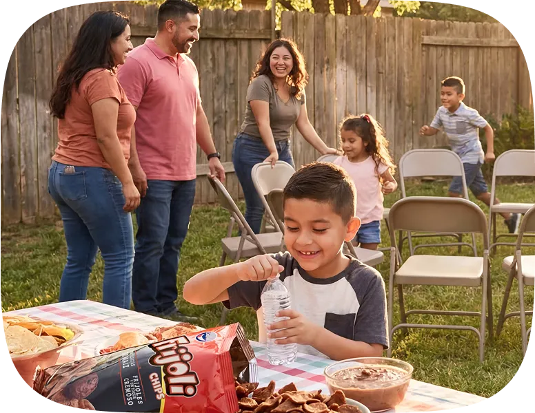 Smiling young boy opening a water bottle at a picnic table with snacks, while a family sets up folding chairs in a backyard.