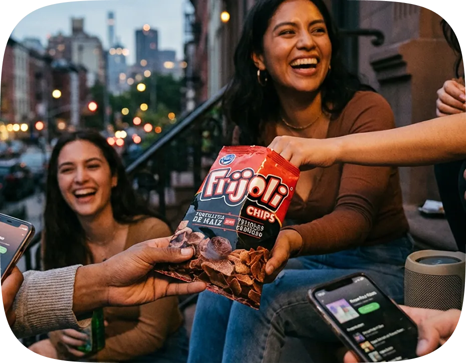 A group of friends laughing and sharing a bag of Frijoli chips on a city street in the evening.