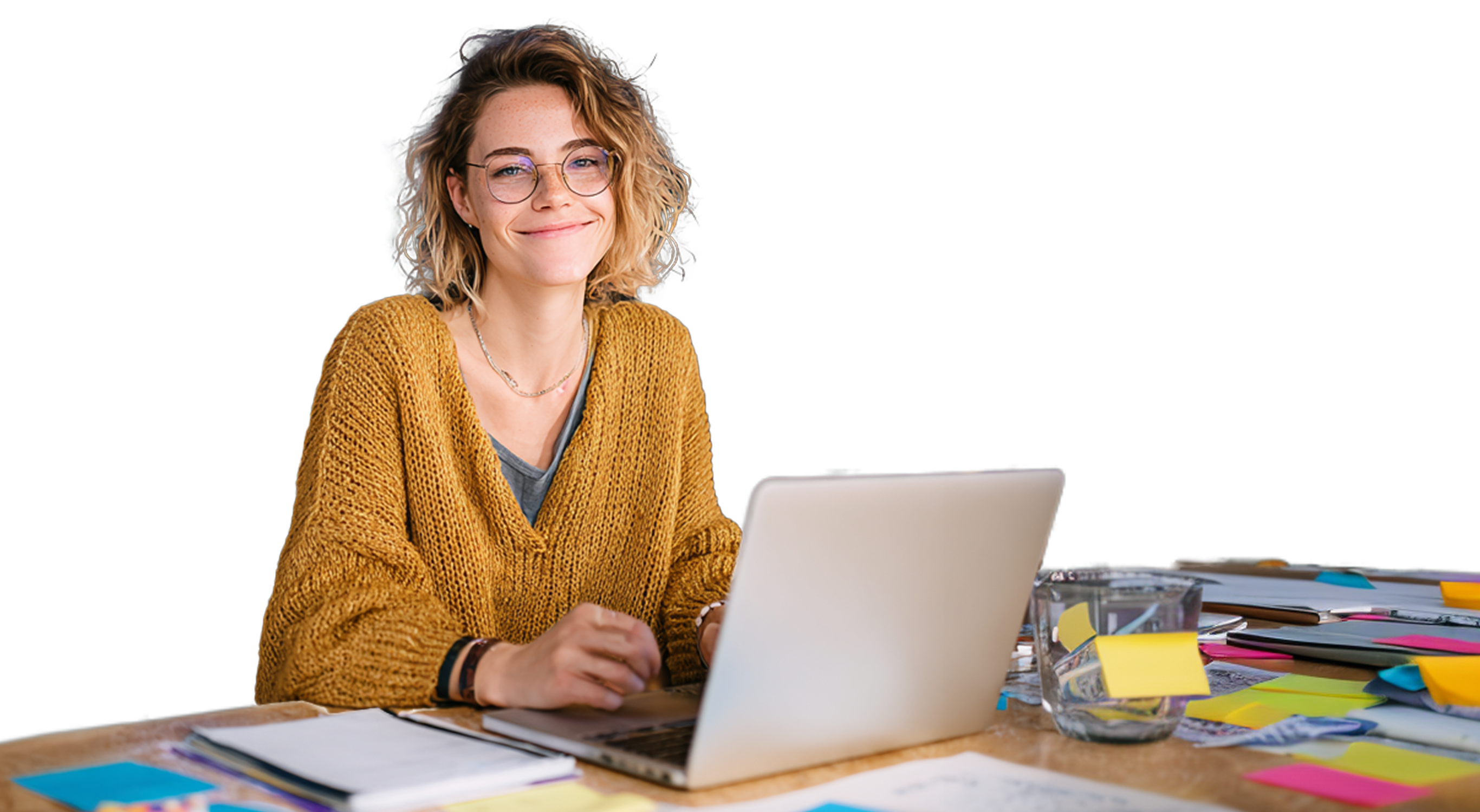 Smiling young woman with curly hair and glasses working on a laptop at a cluttered desk with sticky notes and papers.