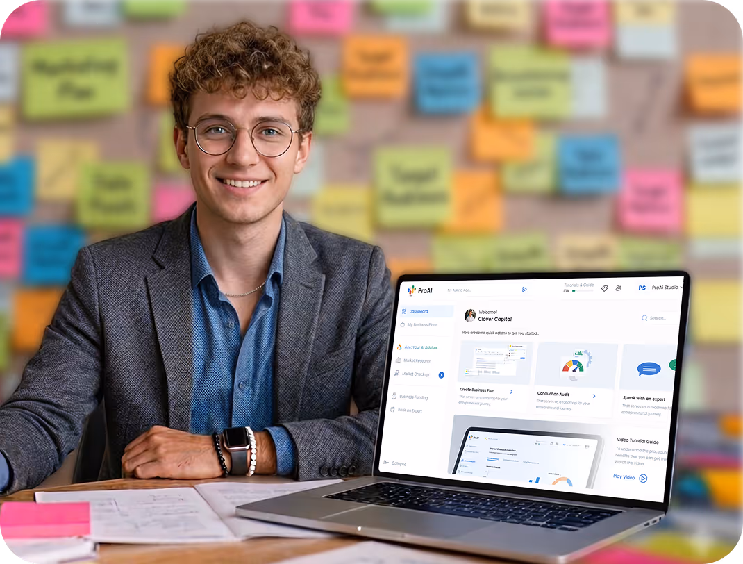 Smiling young man with curly hair and glasses sitting at a table with papers and a laptop displaying a business plan software interface.