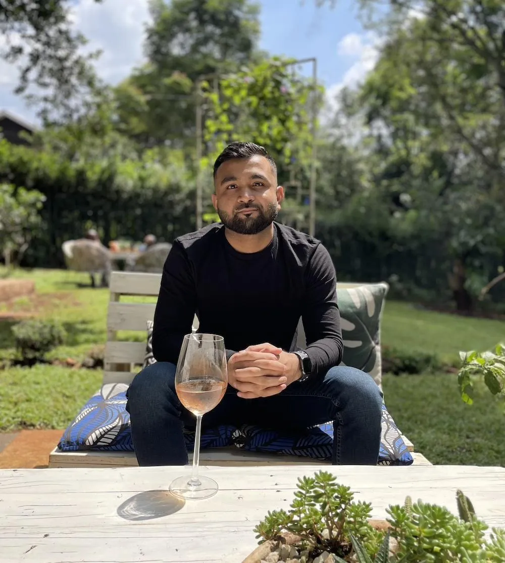Smiling young person with shoulder-length brown hair wearing a white shirt and dark blazer against a solid dark blue background.