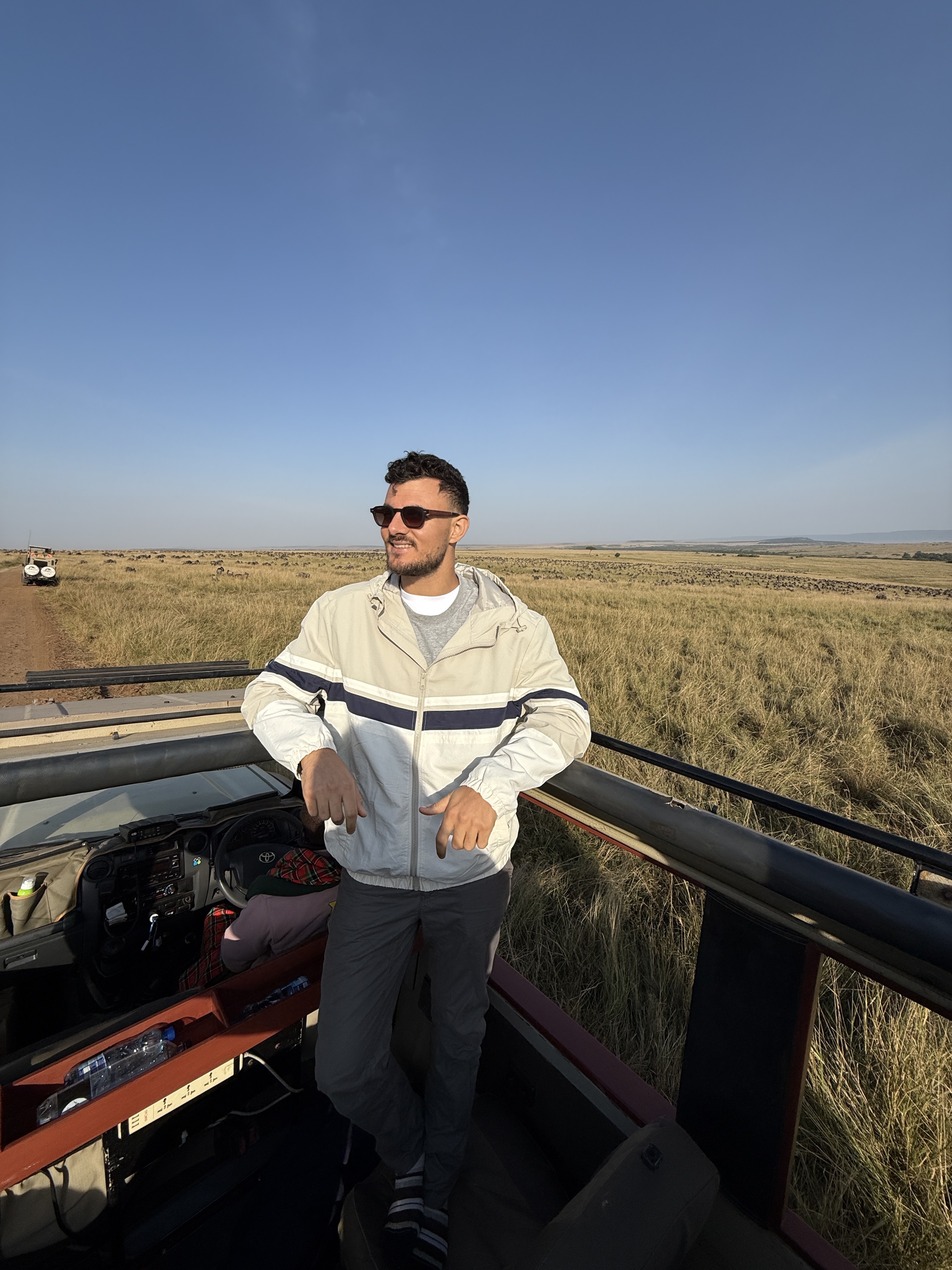 Smiling young person with shoulder-length brown hair wearing a white shirt and dark blazer against a solid dark blue background.