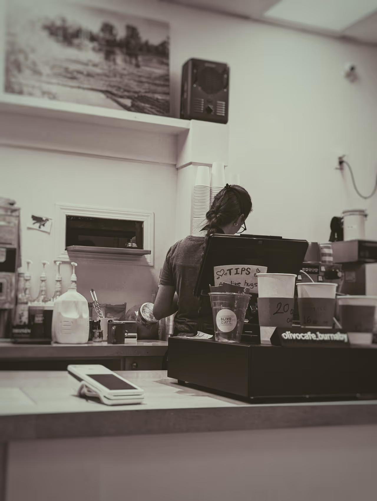 A barista is seen making a drink behind the till at a cafe with a tip jar and products on the counter.