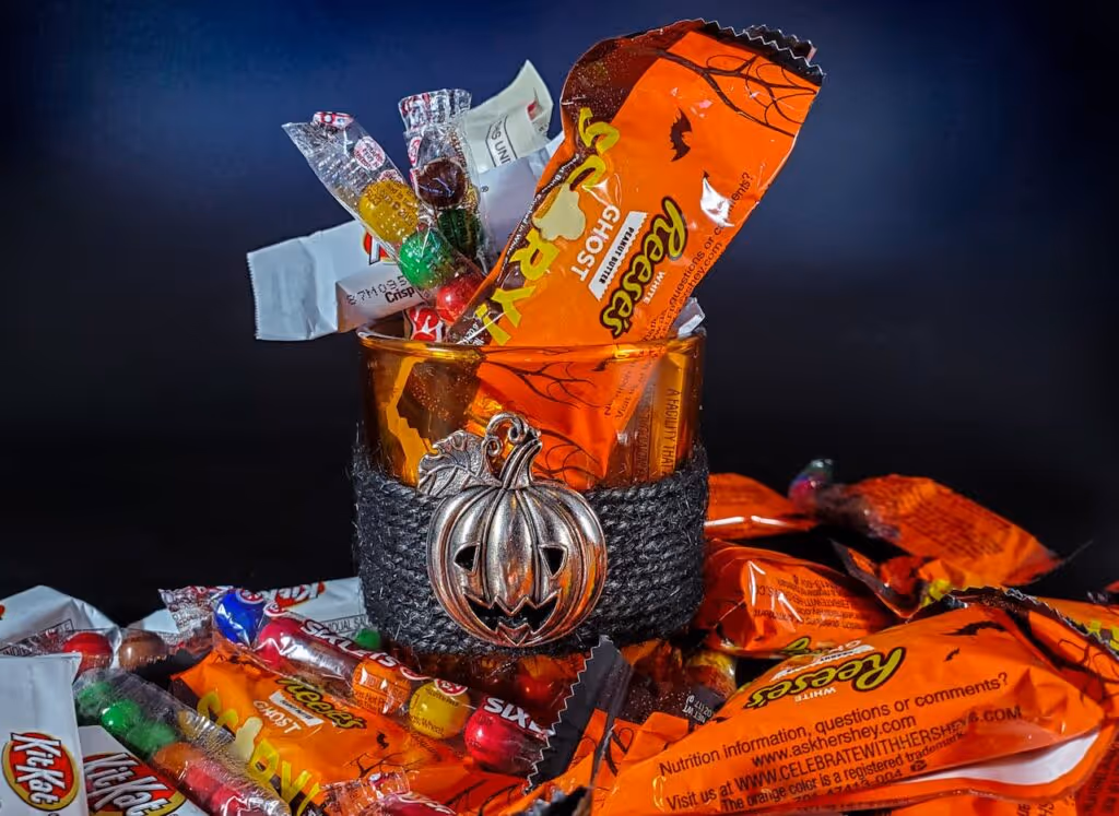 A glass filled with assorted Halloween candies, including Reese's and Kit-Kat, surrounded by more candy and a pumpkin charm.