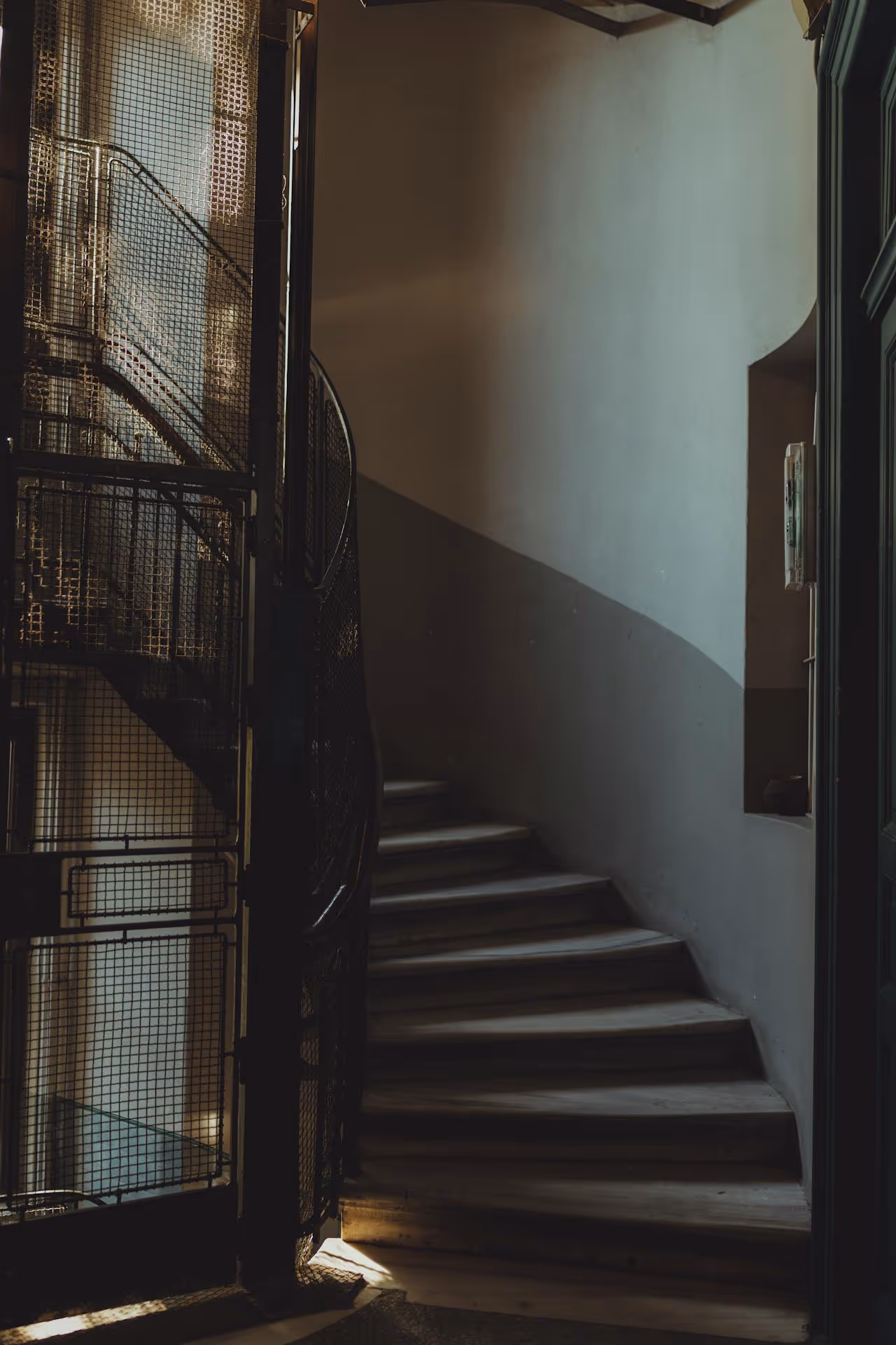 A dimly lit staircase with a spiral metal railing, leading up to a softly illuminated area. The walls are painted in two tones.
