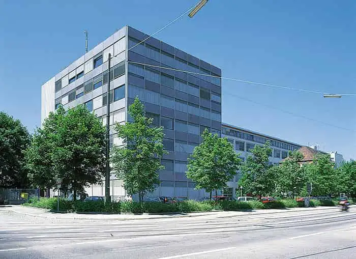 Modernes Bürogebäude mit Glasfassade an einer Straße mit Bäumen und parkenden Autos.