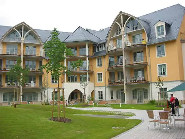 Großes Mehrfamilienwohnhaus mit gelber Fassade, Erkern und Balkonen vor gepflegtem Rasen mit Bäumen und Sitzbänken.