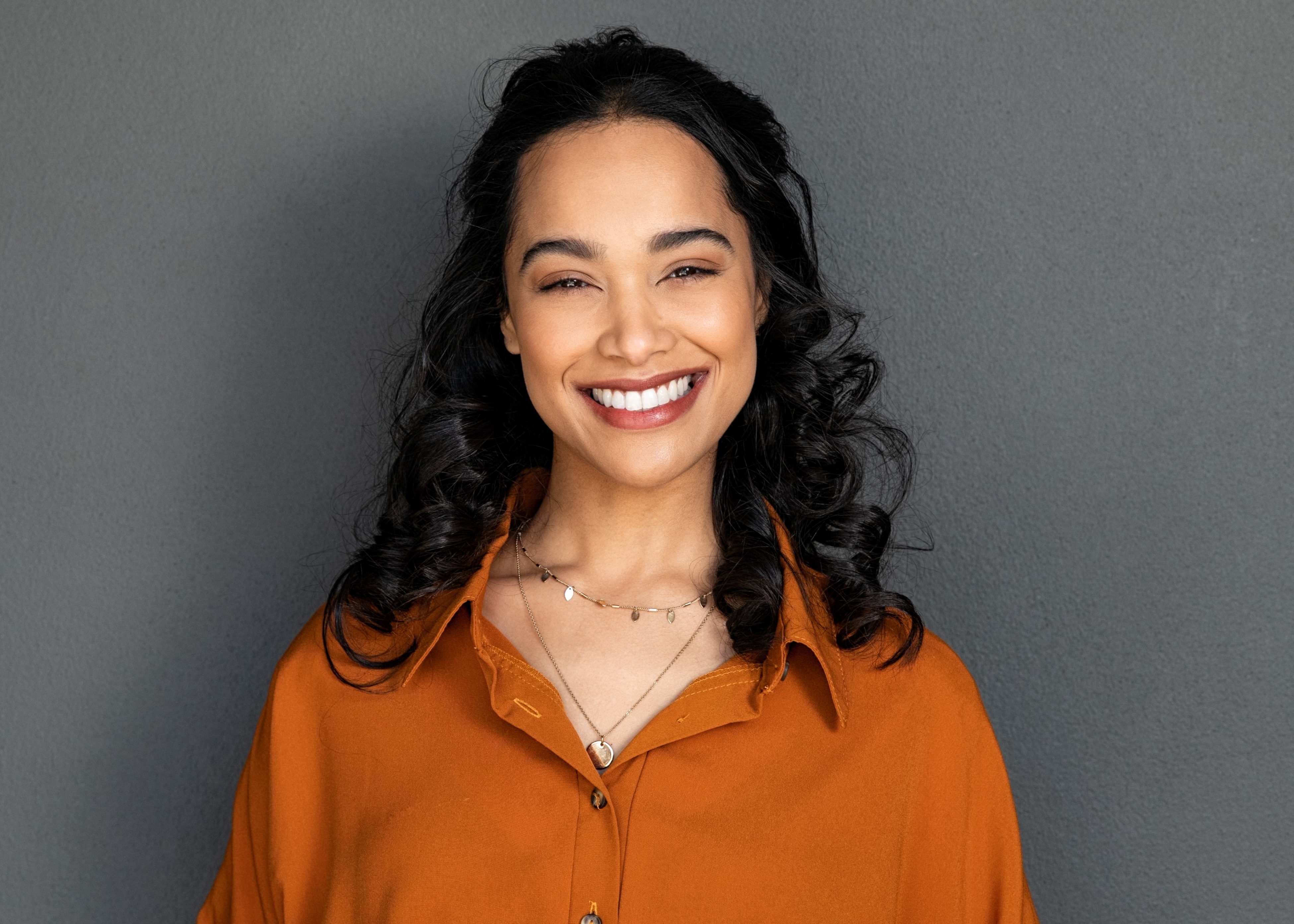 Smiling woman with curly dark hair wearing an orange shirt and two necklaces against a gray wall.