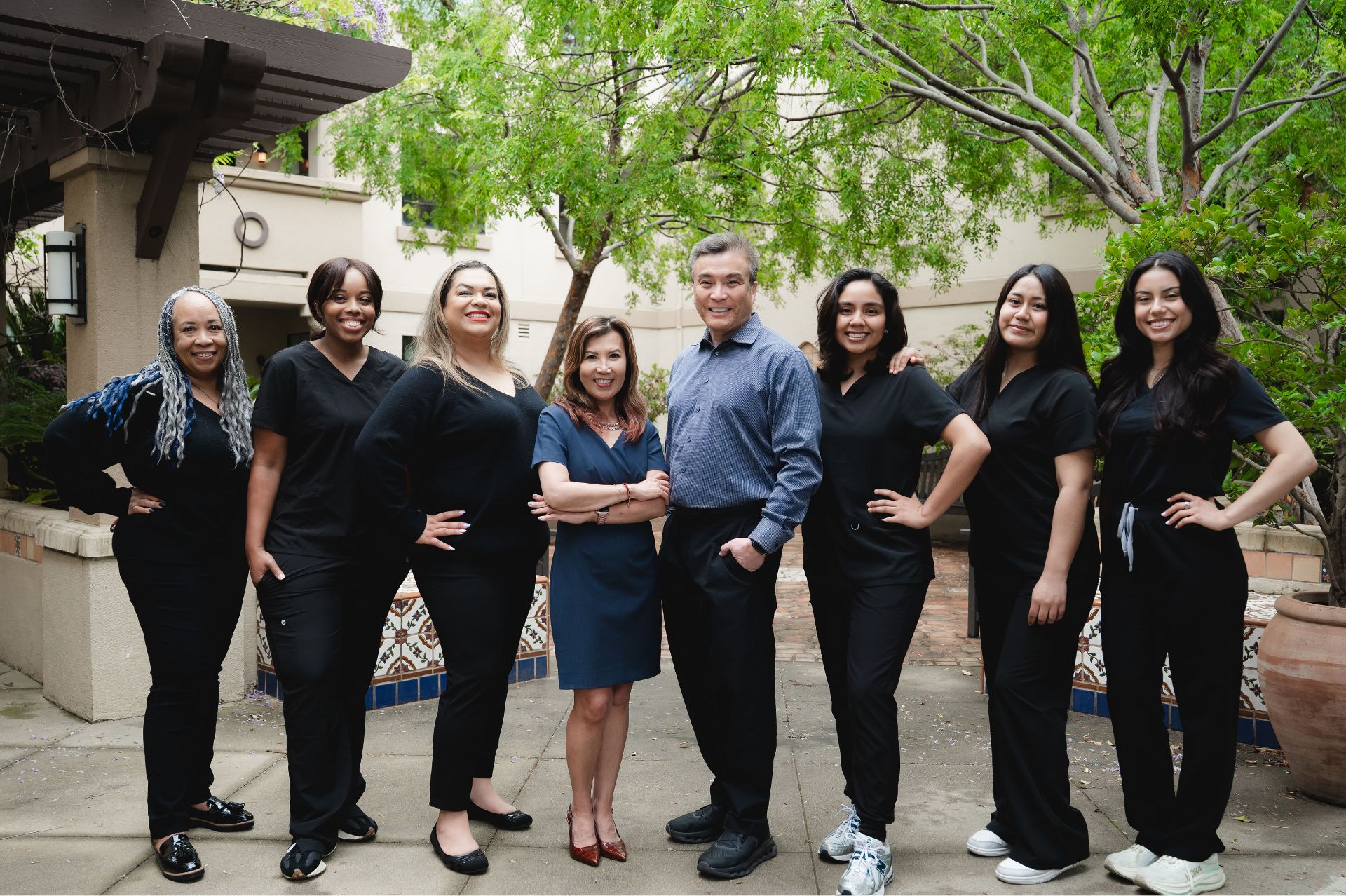 Group of eight diverse healthcare professionals standing outside under green trees, smiling at camera.