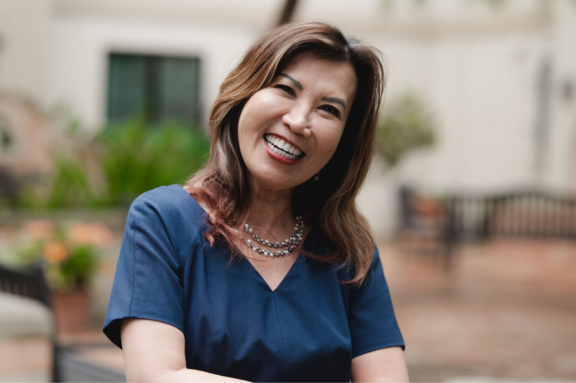 Smiling woman with brown hair wearing a blue top and pearl necklace outdoors near benches.