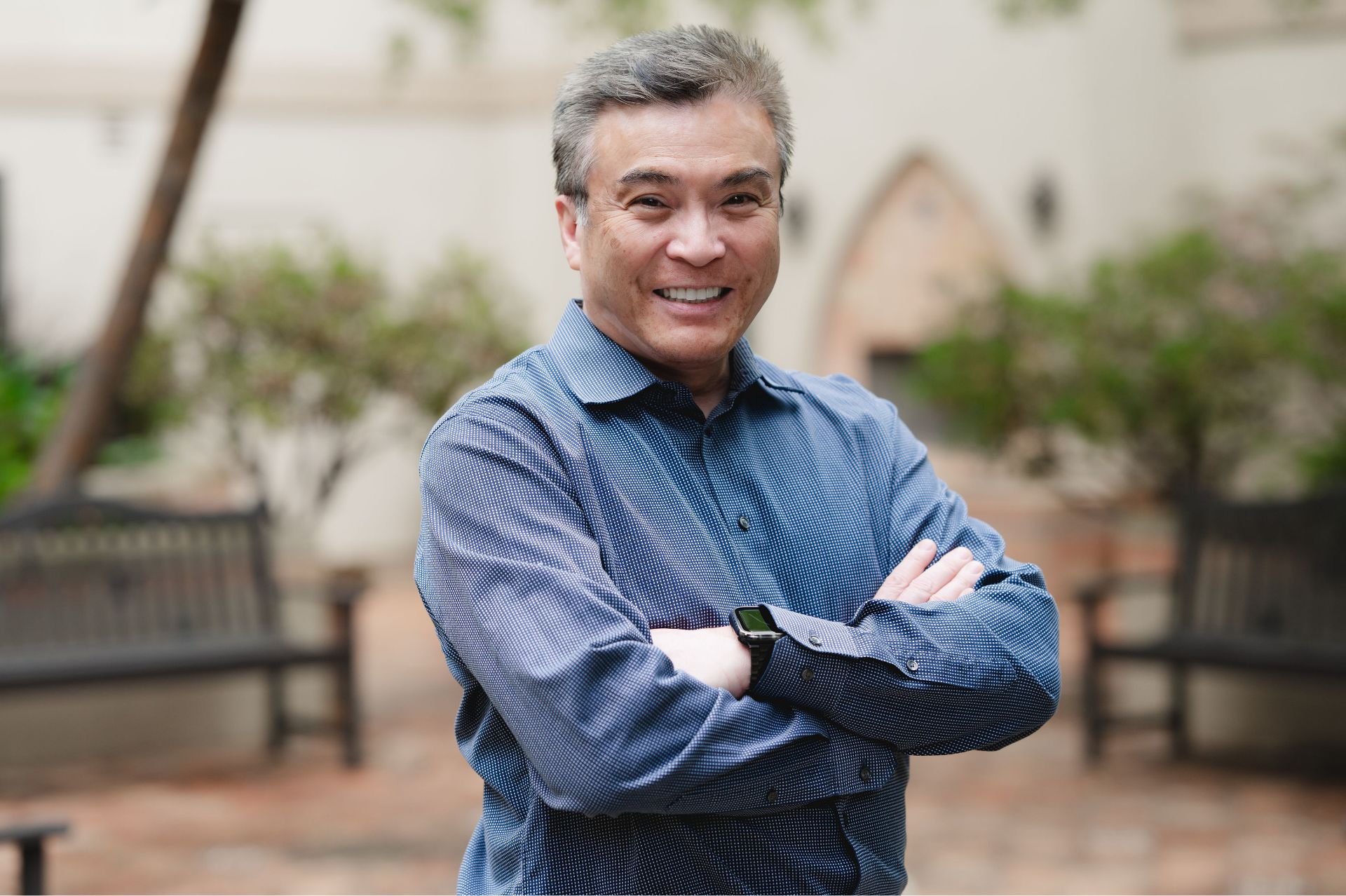 Smiling middle-aged man in a blue buttoned shirt with arms crossed, outdoors near benches and plants.