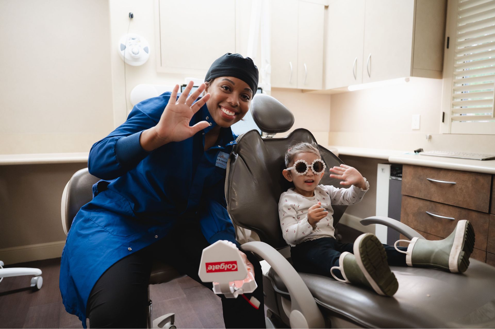 Smiling dentist shows teeth model while a child with sunglasses waves sitting in dental chair.