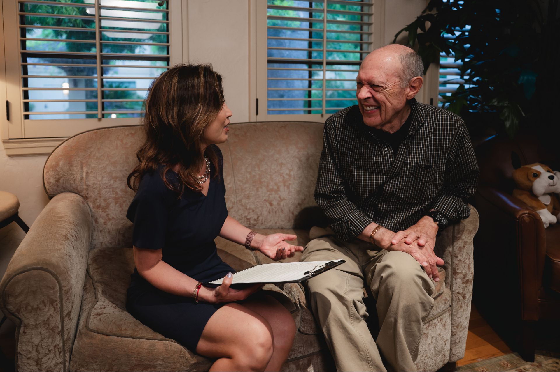 Woman holding clipboard talking with elderly man on a couch in a cozy room with shuttered windows.