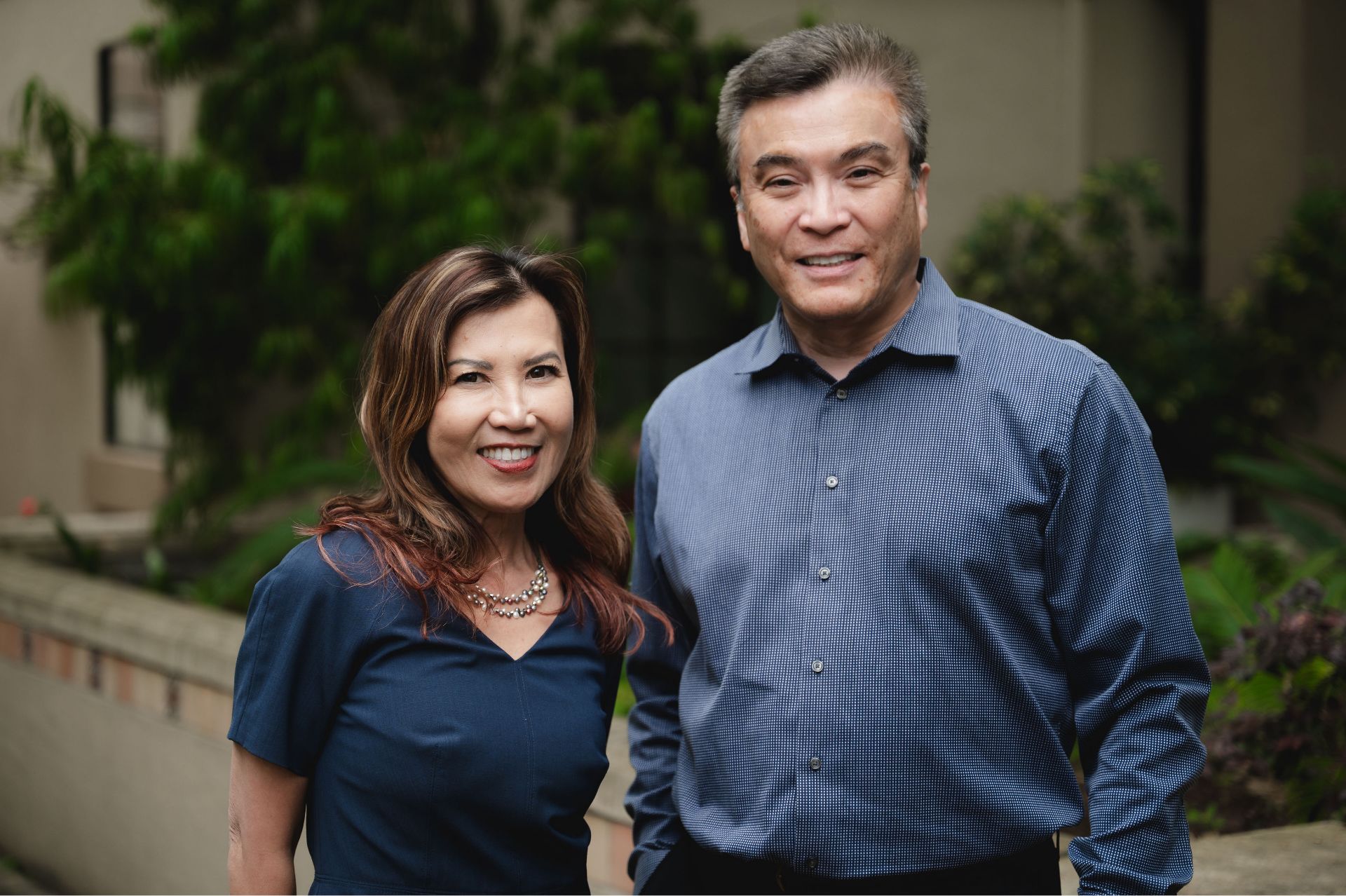Smiling middle-aged man and woman standing outdoors with greenery in the background.