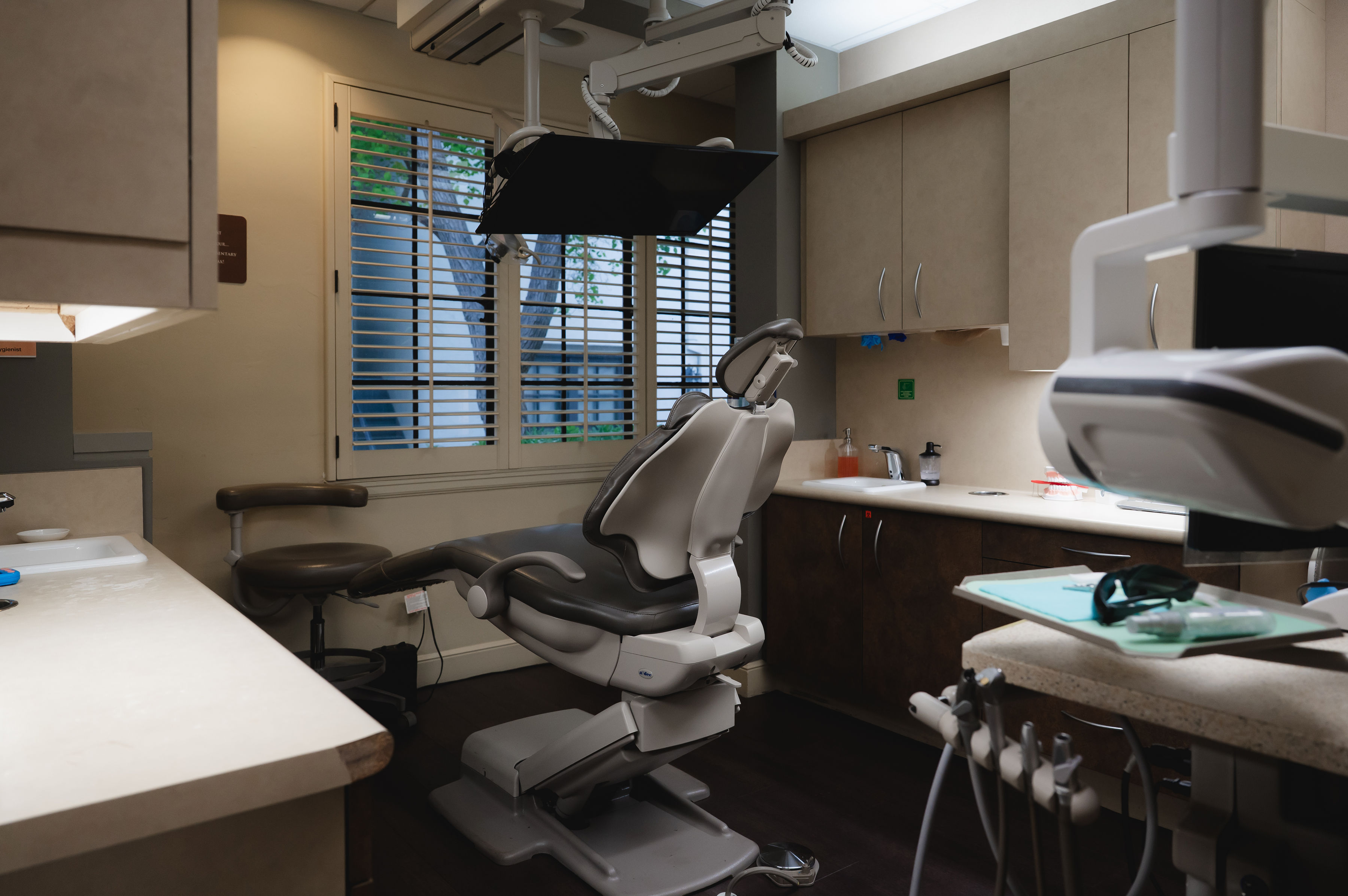 Empty dental exam room with chair, overhead light, and dental tools near a window with blinds.