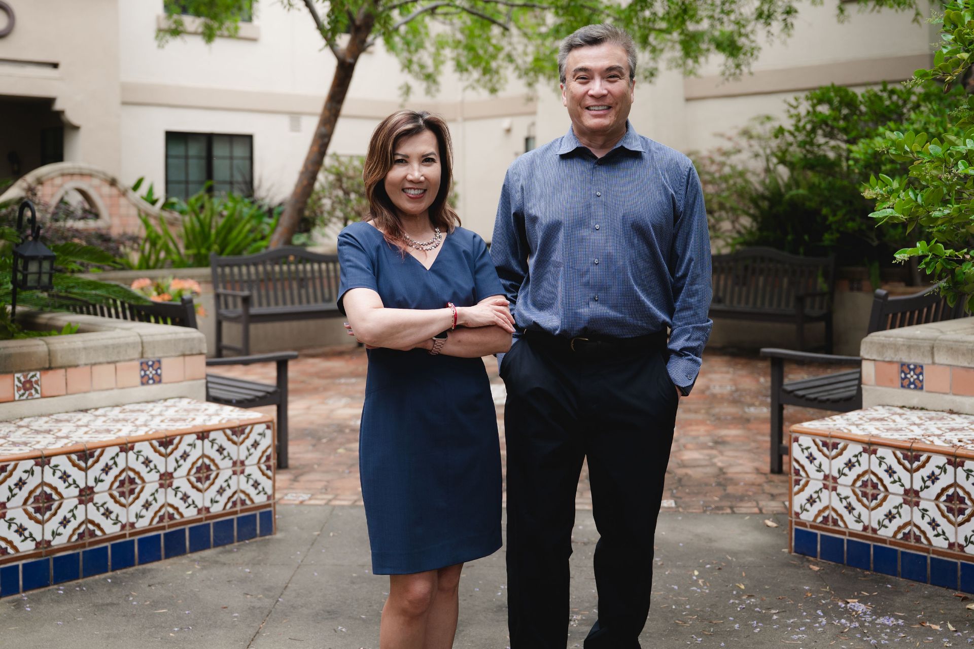 Smiling man and woman standing side by side in a tiled courtyard with benches and plants.