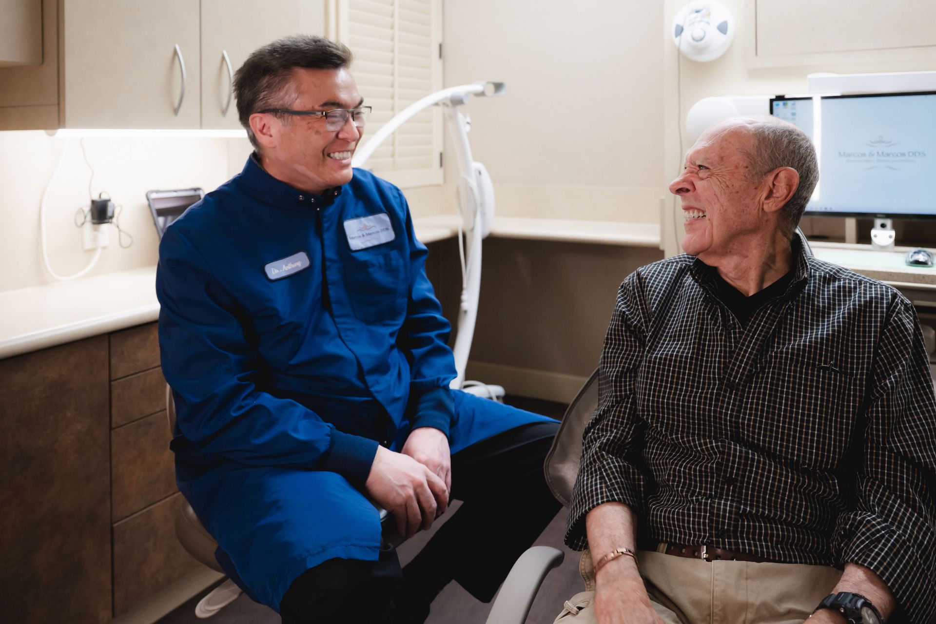Dentist in blue coat smiling and talking with elderly male patient in dental office.