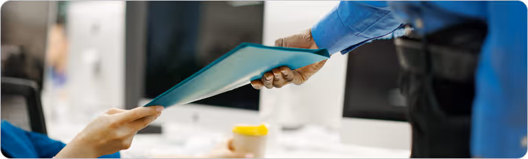 Two office workers passing a blue file folder to each other in a professional workspace.