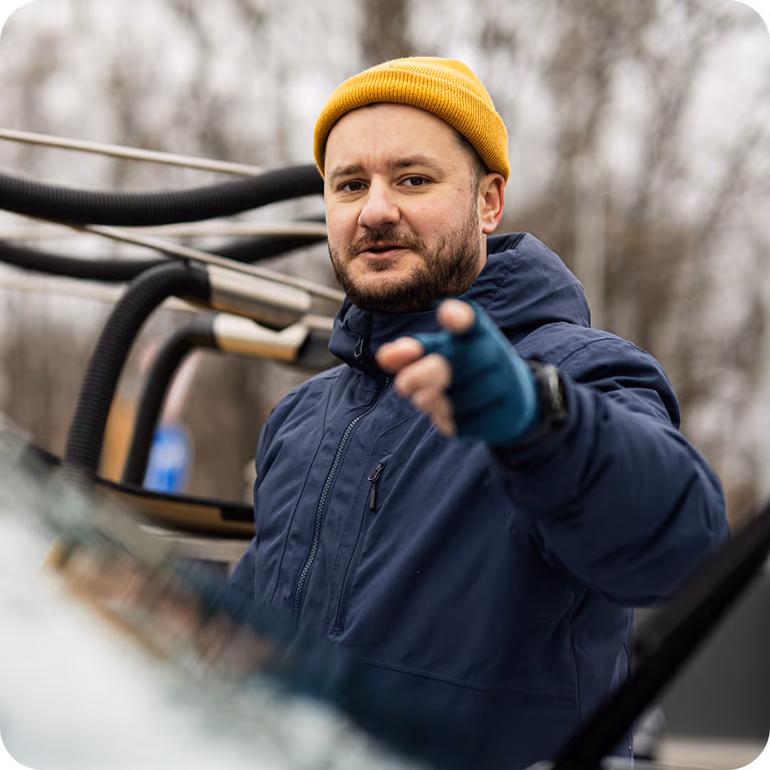 A man in a blue winter jacket and yellow beanie pointing forward while standing outdoors near a vehicle with roof equipment.