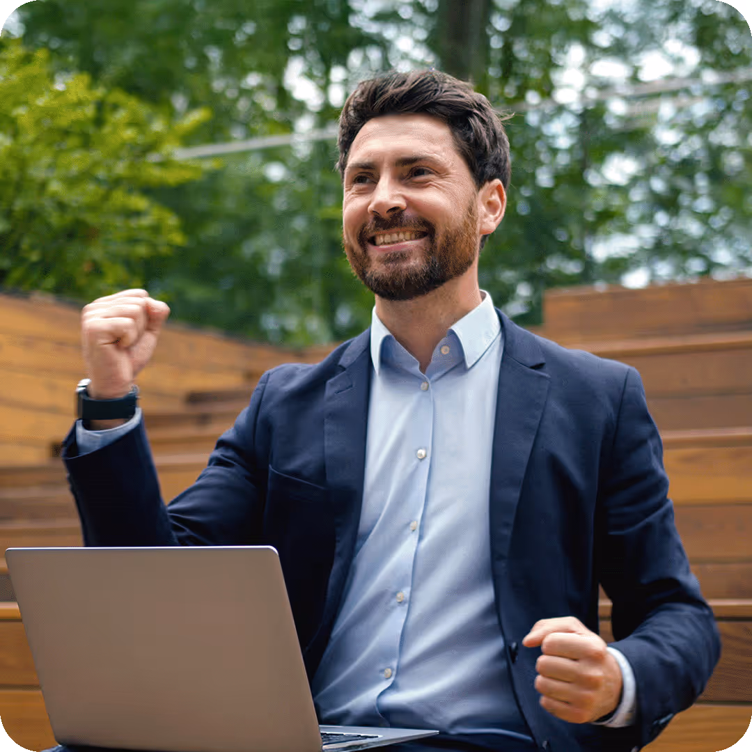 A businessman sitting outdoors with a laptop, cheering with a clenched fist to celebrate a successful project or career milestone.