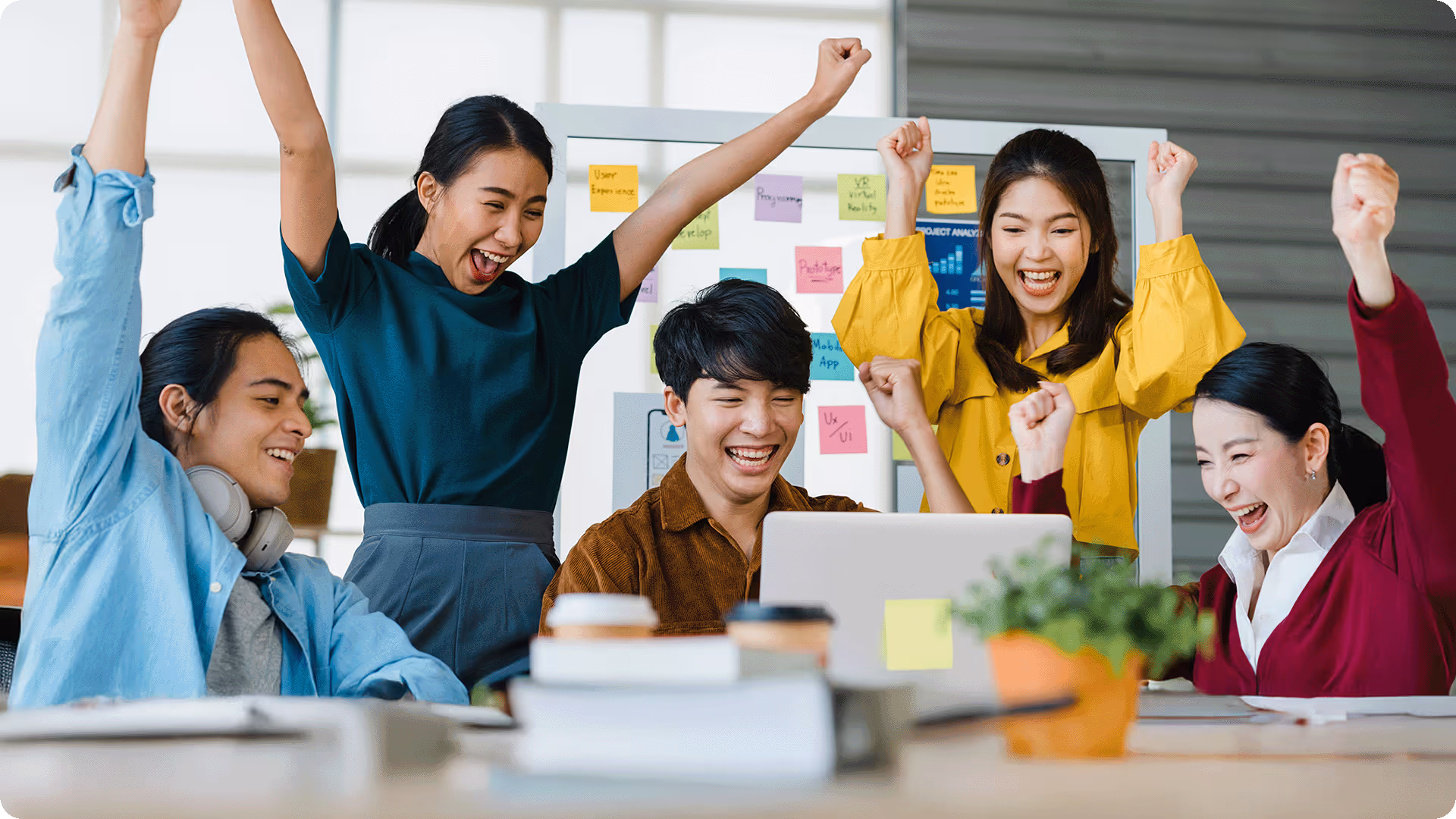 A diverse team of young professionals cheering and celebrating a win while looking at a laptop in a creative office.