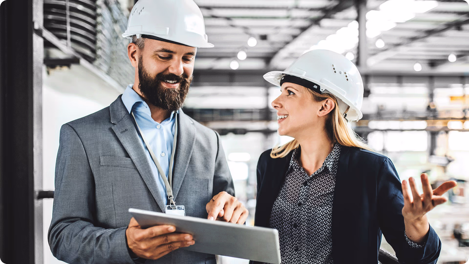 A male and female facility manager wearing white hard hats and professional attire discussing data on a digital tablet at a worksite.