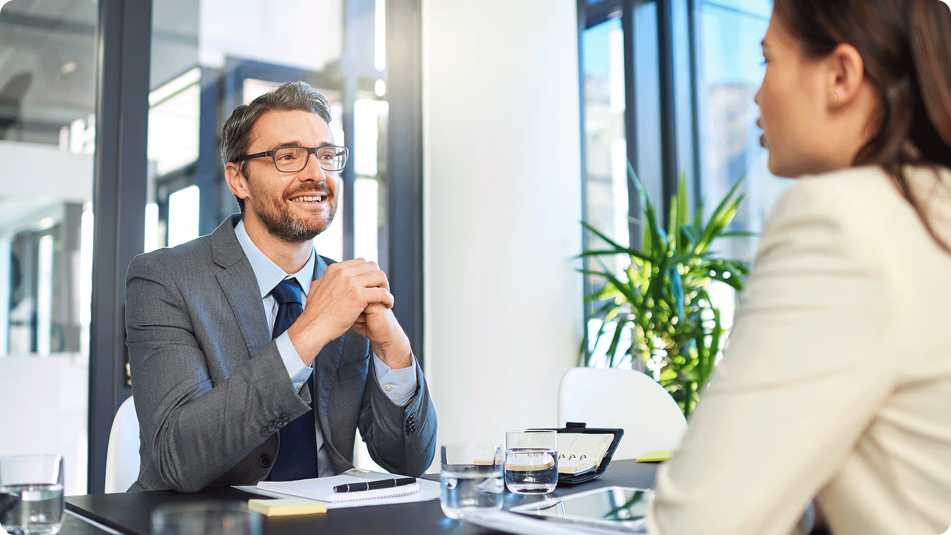 A male hiring manager in a grey suit smiling during a face-to-face job interview with a candidate in a bright office.